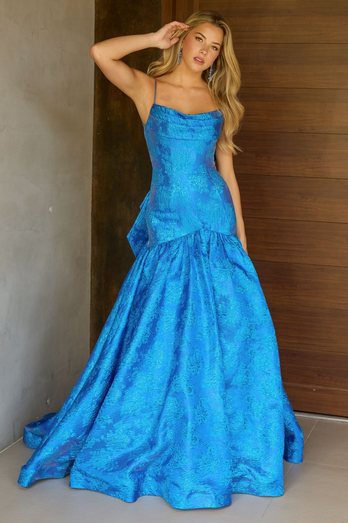 A woman stands indoors by a wooden door and gray wall, wearing the Ava Presley 26419 Drop-Waist Long Print Prom Gown—a floor-length, bright blue textured dress with a fitted bodice and flared skirt.
